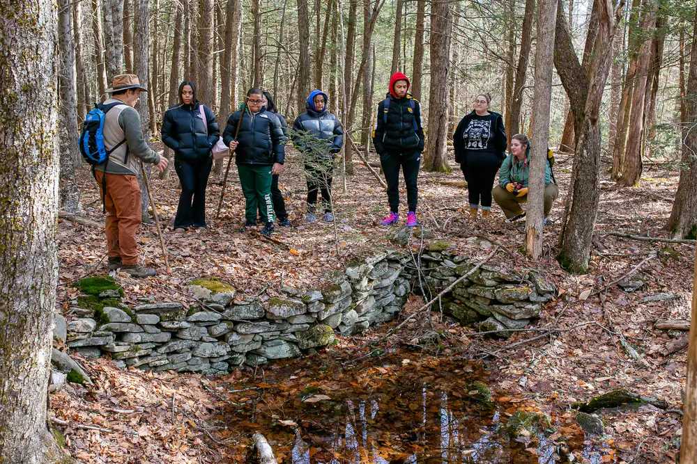A guide and a group of hikers standing along the mill's low stone foundation in bare winter forest